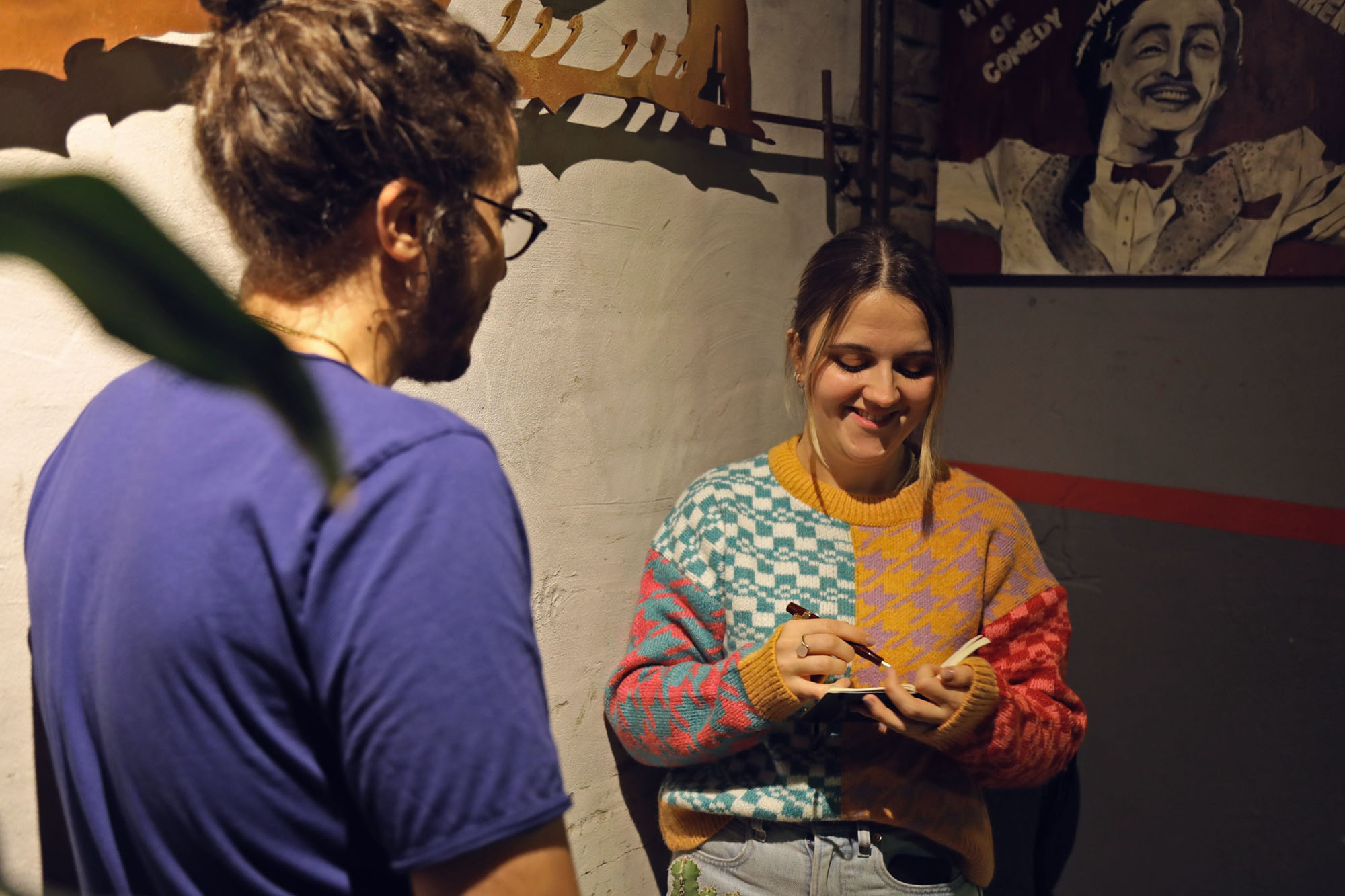 Photo: Sümeyra Güneş (right), a woman with dark brown hair and wearing a colorful sweater smiles as she takes notes as she interviews comedian Murat Gencoglu, a man facing away from the camera wearing a purple shirt and glasses.