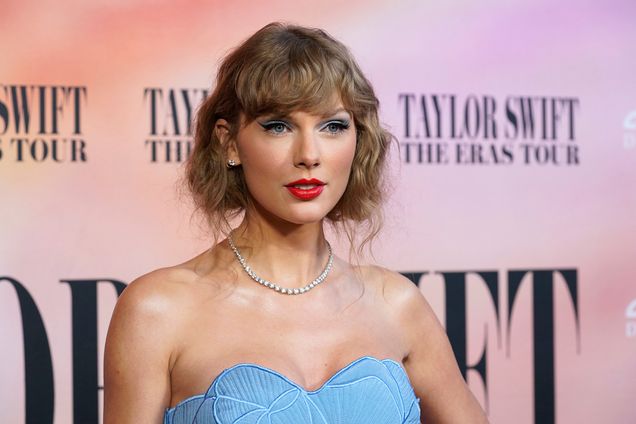 Photo: Taylor Swift, a white woman wearing a light blue halter top dress, smiles and poses in front of a backdrop that reads "Taylor Swift, The Eras Tour".