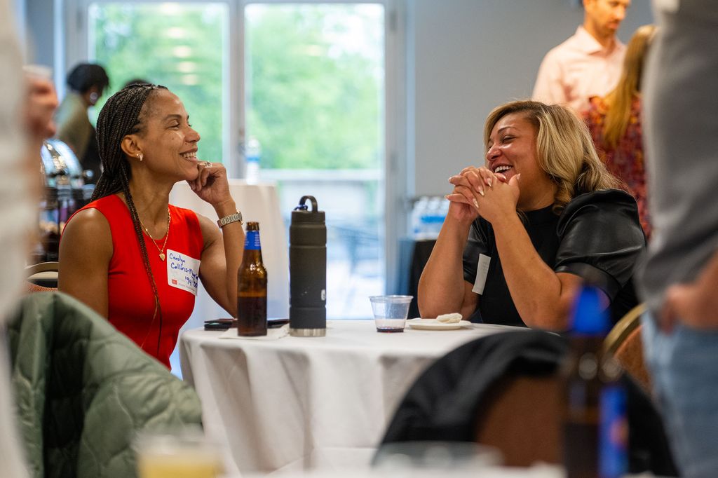 Photo: Two women sit and mingle at a white table. On the left, a light skinned Black woman with medium box braids and wearing a red dress smiles as she listens to the woman sitting across from her, a light-skinned Black woman with blonde hair and wearing a black blouse.