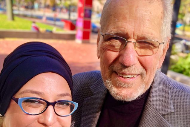 Photo: A man and woman stand side by side with smiles on their face, both are wearing glasses and it is sunny outside