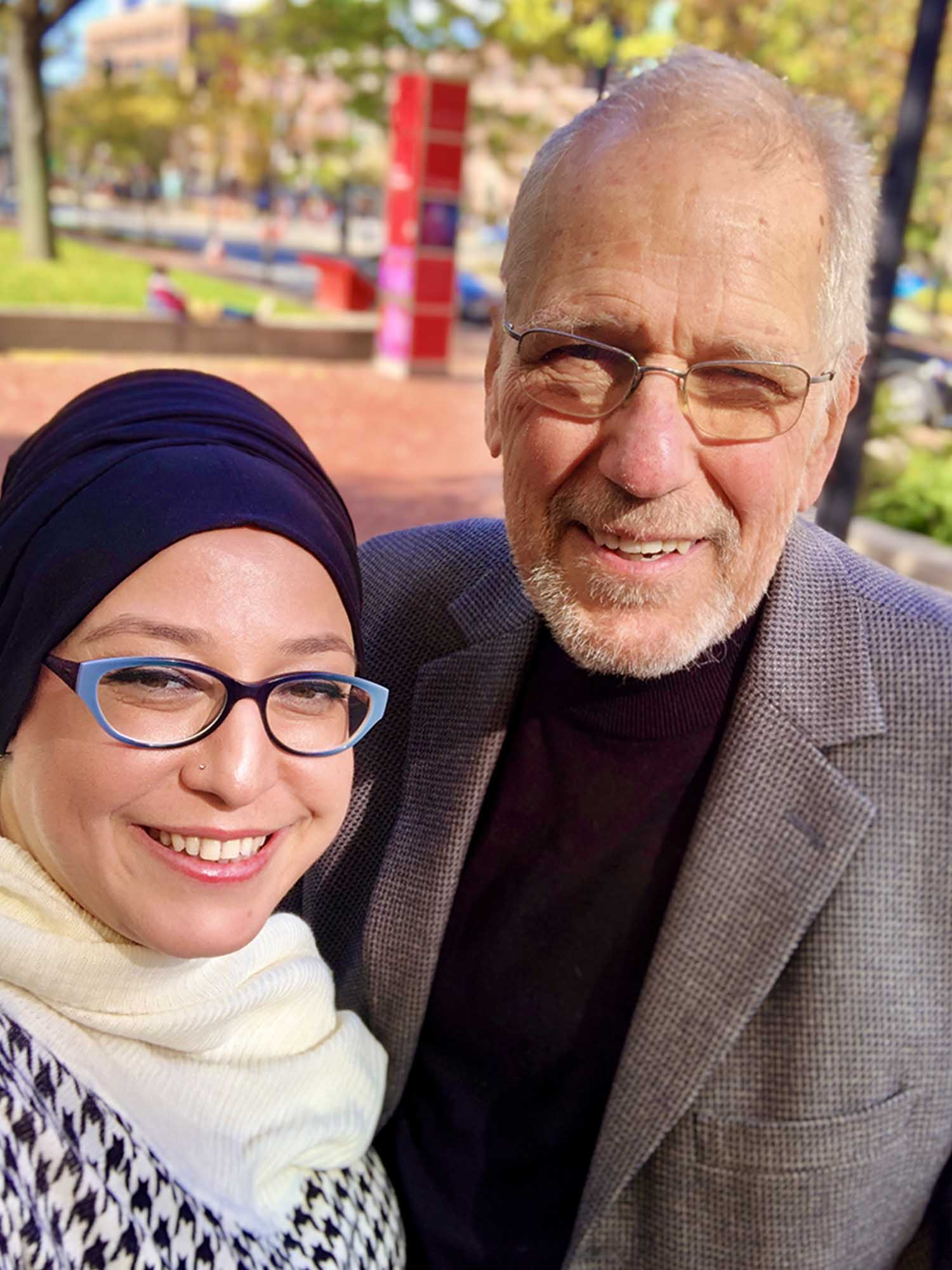 Photo: A man and woman stand side by side with smiles on their face, both are wearing glasses and it is sunny outside