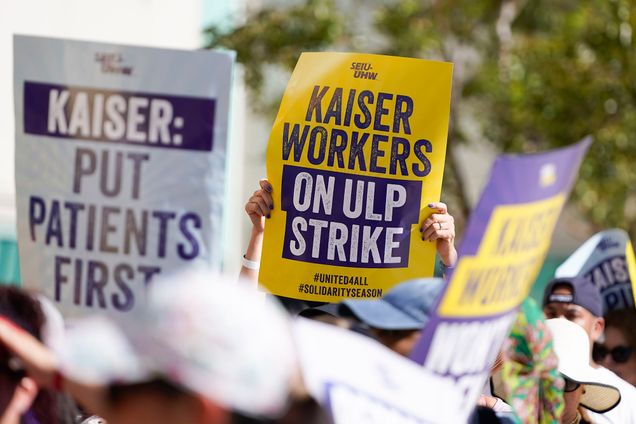 Photo: People hold up large yellow and purple signs in support of the ULP Strike. Photo highlights one sign that reads "Kaiser Workers on ULP Strike".