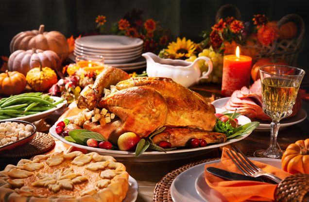 This is a stock photo from Canva of a Thanksgiving dinner spread. There is a cooked turkey in the middle with pies, vegetables, and other foods spread out around it. There are also candles, pumpkins and flowers to create a fall theme.