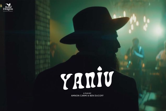 Image: Movie poster for "Yaniv". An Israeli man wearing a large hat is shown in shadow looking to the right as he enters a low-lit, spooky room. Large white letters in bottom center read "Yaniv". Small logo in top left reads "Roof Dragon Entertainment".