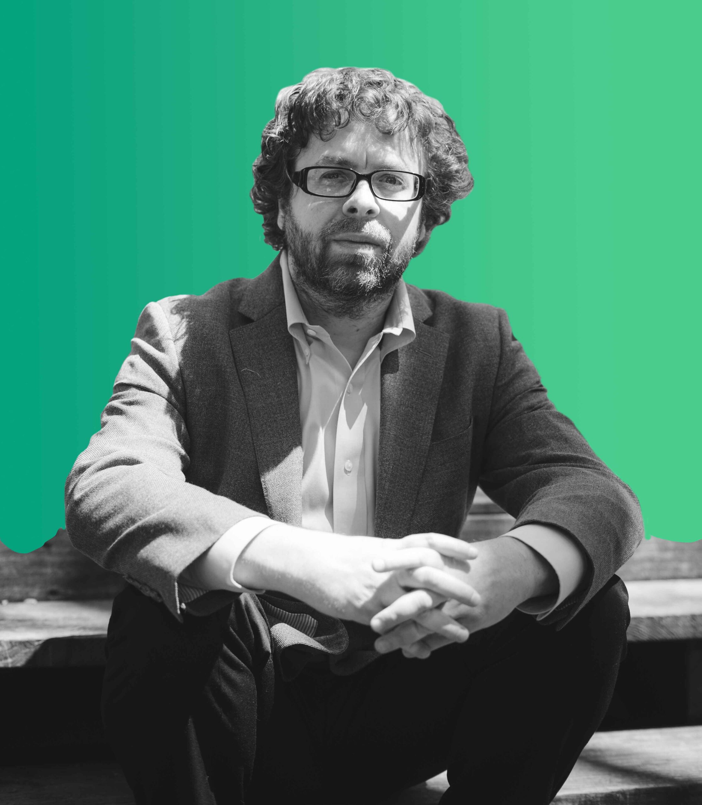 Photo: A black and white cut out of Elon Green, a man with short, curly hair wearing glasses and a suit, clasping his hands as he sits and stares at the camera. A green gradient is the background.