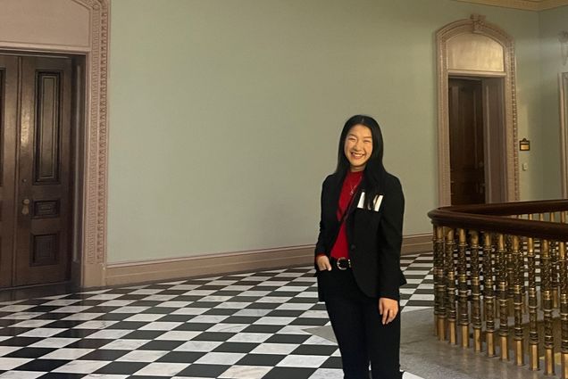 Photo: A smiling woman in a suit and blazer stands in a checkered floor in an ornate room near a wooden carved railing
