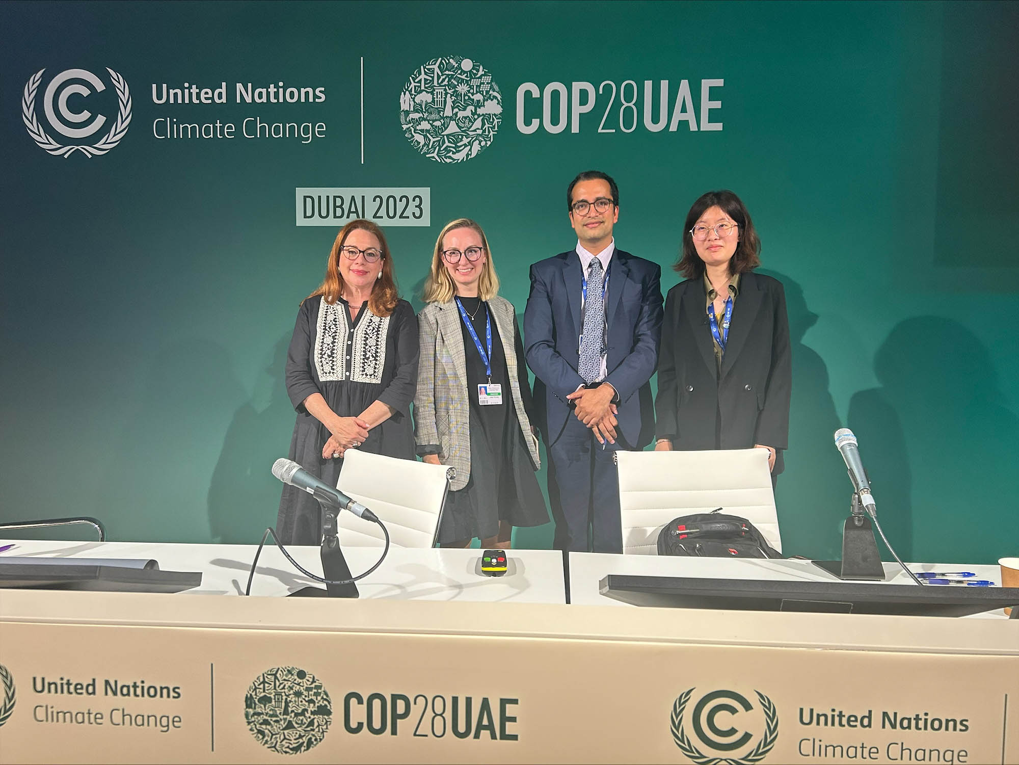 Photo: Four people wearing business casual attire pose in front of a large teal backdrop that reads "COP28UAE". They stand behind a long table fitted with various chairs ands microphones. The tables also reads "COP28UAE".