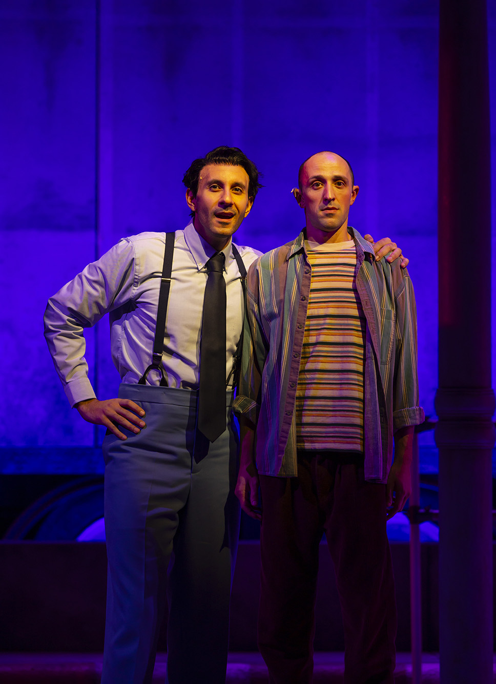 Photo: Band member Haled, a white man with curly hair and wearing a white collared shirt, black tie, and grey slacks and suspenders looks to the audience as he gives advice to the man to his right. He has his arm around a balding man wearing a yellow and black striped shirt and colorful, unbuttoned collared shirt. They both stand on stage and face the audience as purple and blue lights light up the stage.