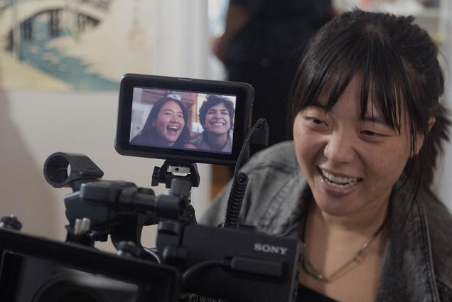 Photo: A young Asian woman is shown filming using a large black film camera. The small display on the camera shows two young adults in the midst of being filmed for a scene.