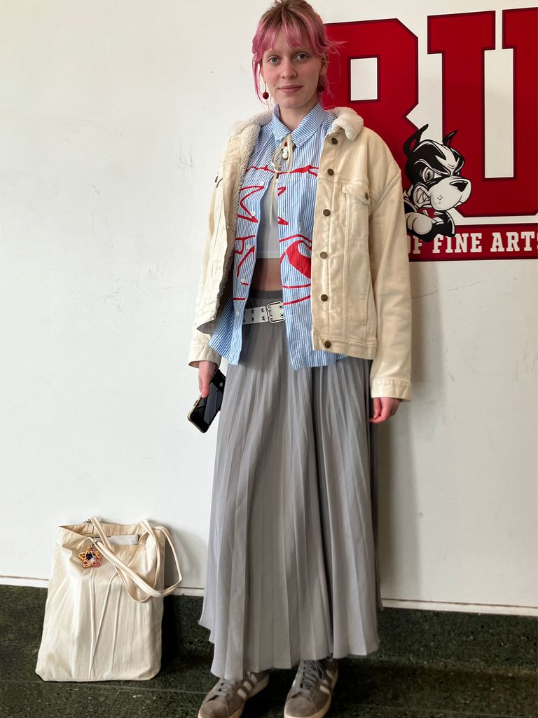 Photo: A college student with short blonde hair wearing a long gray dress and a white jacket. They are standing in front of a red BU logo.