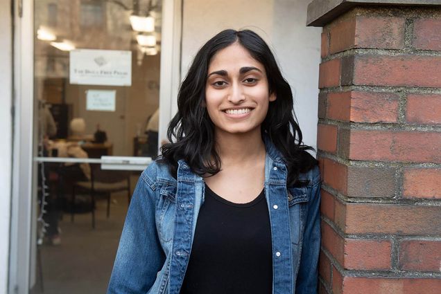 Photo: Madhri Yehiya, a young South Asian woman with curled black hair and wearing a black shirt and jean jacket smiles and poses outside a clear door of a brick building.