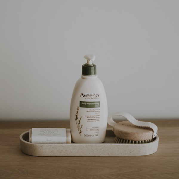 Photo: A small display of shower skincare is shown on a wooden table. A bar of soap, large Aveeno lotion, and small dry scrub brush are shown in a small clay tray.