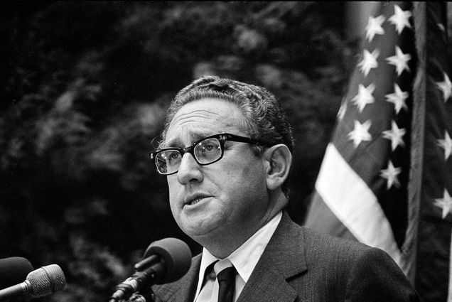 Photo: A black and white photo of Henry Kissinger when he was Secretary of State. He stands at a podium, talking to a crowd in front of a flag of the United States.