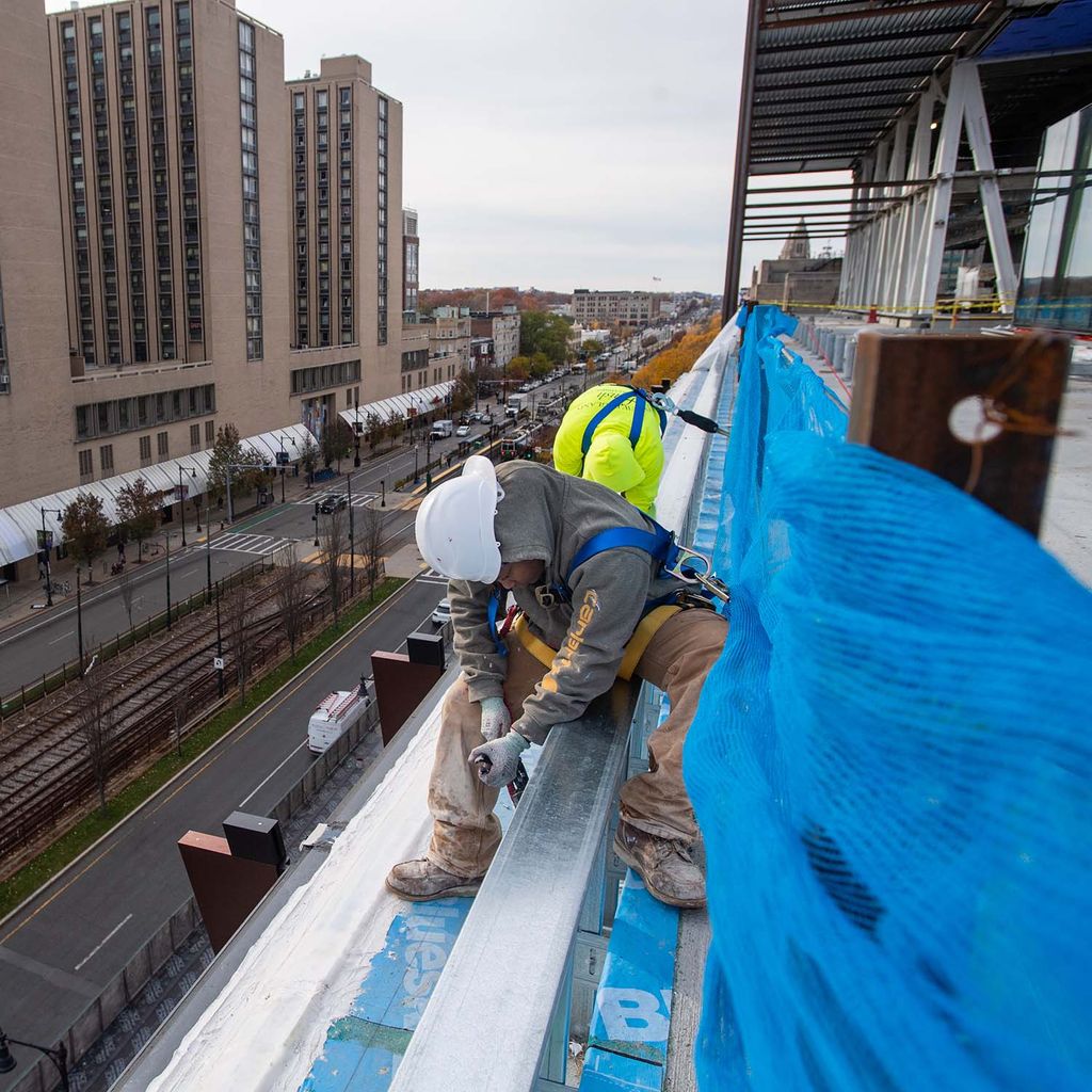 Photo of a construction worker sitting at the edge of a floor of the data sciences building and removing a blue tarp from the ground. Comm Ave is seen below him.