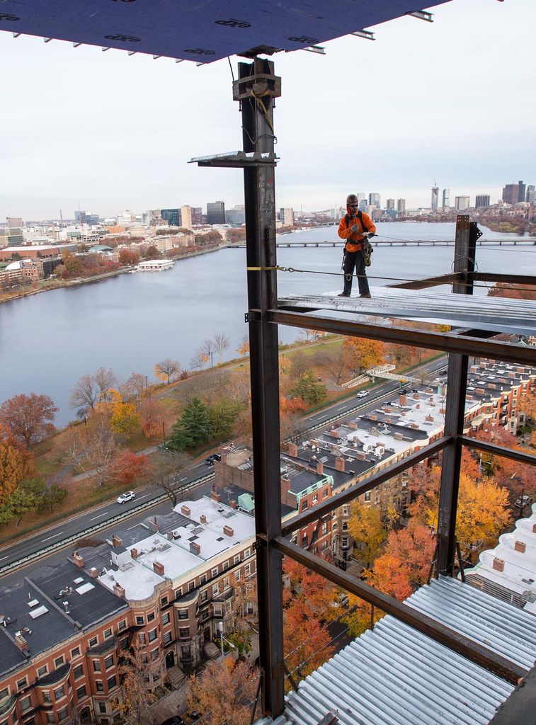 A construction worker dressed in orange looks on as a beam is about to be removed from the side of the data sciences building. In the background, Storrow drive and the charles river are seen, as well as trees with fall-colored leaves.