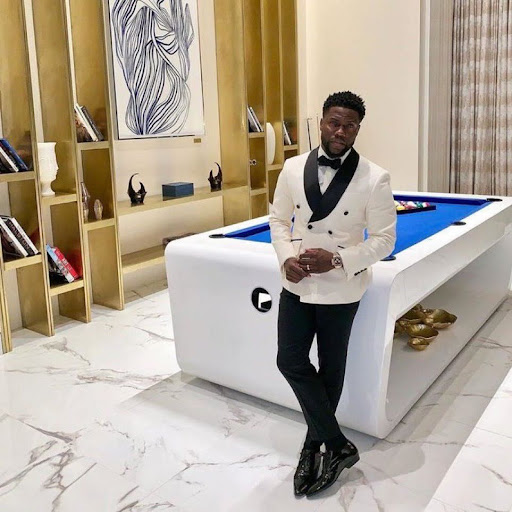 This is a photo of comedian Kevin Hart leaning against a pool table. he is wearing a white and black colored tuxedo with his legs crossed. He is looking at the camera with a straight face.