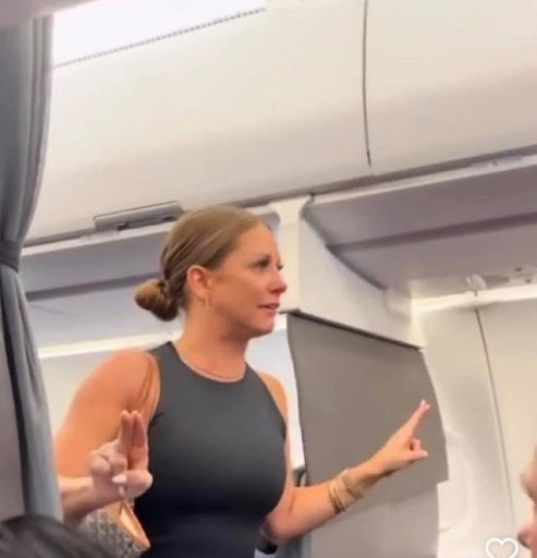 This is a screenshot from a viral video of a woman on an airplane who was claiming to see an extra terrestrial on the plane. She is standing up, pointing her fingers to the side. She has her tied back and it wearing a black tank top.