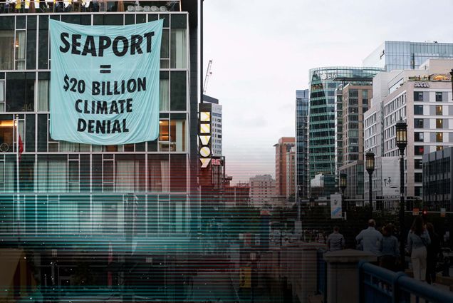 Photo: A large banner on the side of a building in Boston's Seaport reading "Seaport = 20 Billon Dollar Climate Denial" as people walk by in their daily routines
