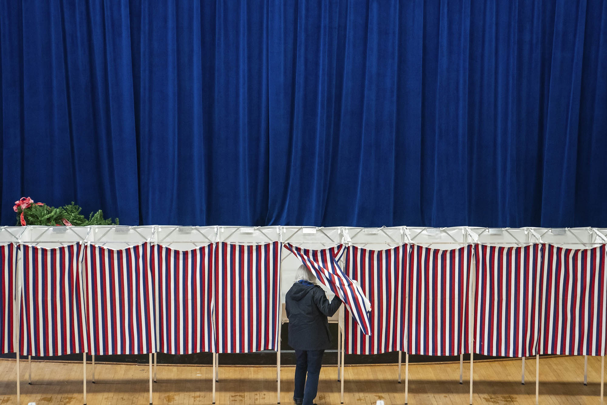 Photo: A generic wide shot of polling booths with red, white, and blue curtains draped in front. In the middle of the photo, a person sneaks into a booth.