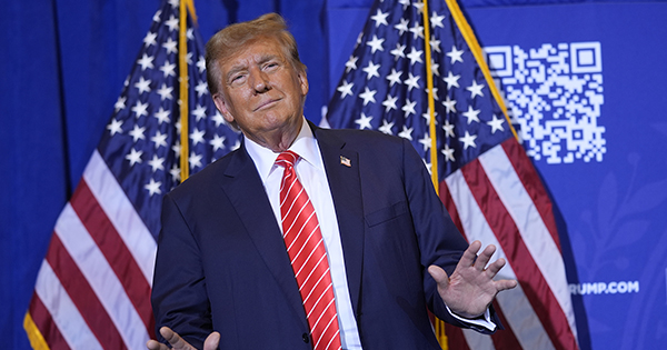 Photo: Donald Trump in a navy suit in front of the american flag. Trump has a smug look on his face and his hands raised.