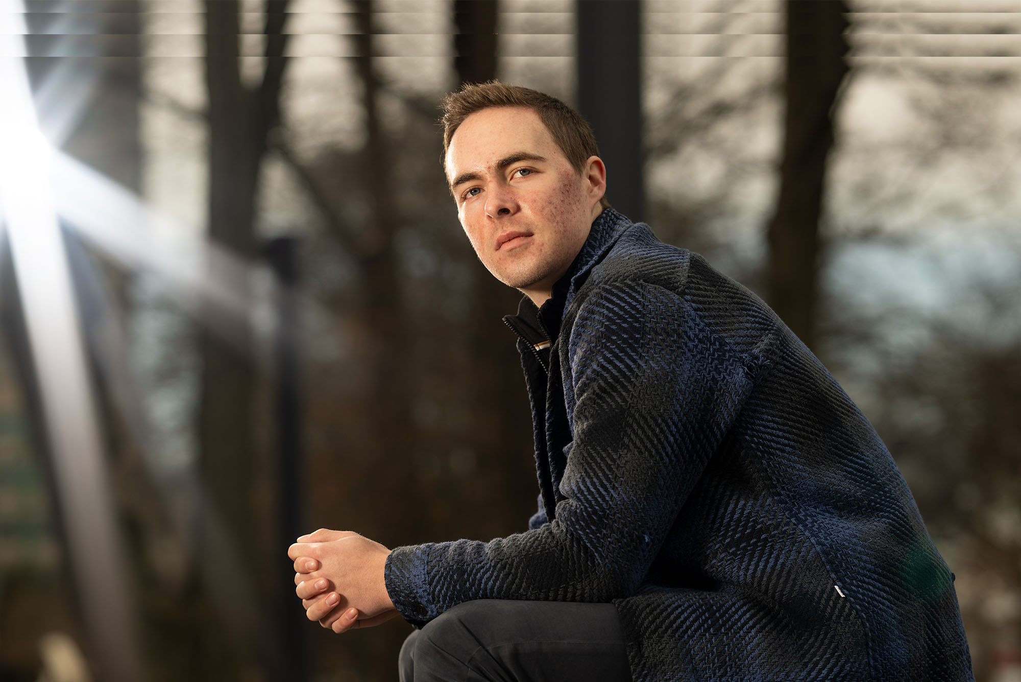 Photo: Cameron Monesmith, a white man with short hair wearing a flannel shirt, sits outside, his left side profile to the camera, hands clasp, posing for the photo.
