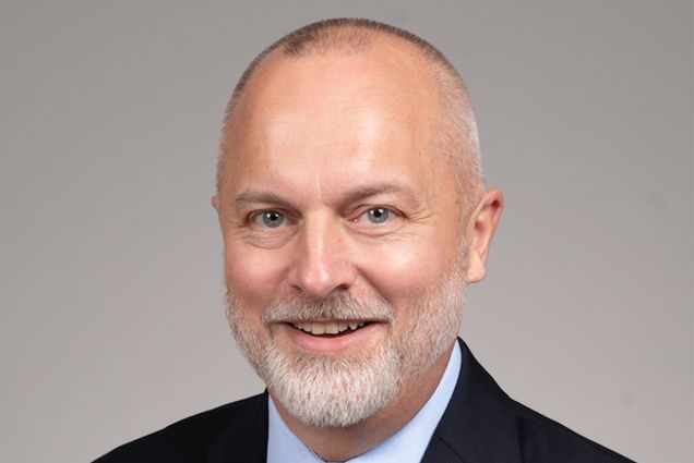 Photo: A white man wearing a suit and golden tie with a soft smile