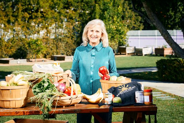 Photo: A white woman with a teal button up and teal slacks poses for a photo. She smiles for the photo with her blonde hair. She poses with her products on a table in front of her.