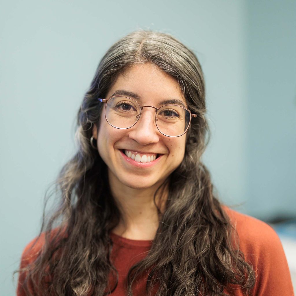 Photo: Headshot of a woman with long curly hair, glasses and a red shirt
