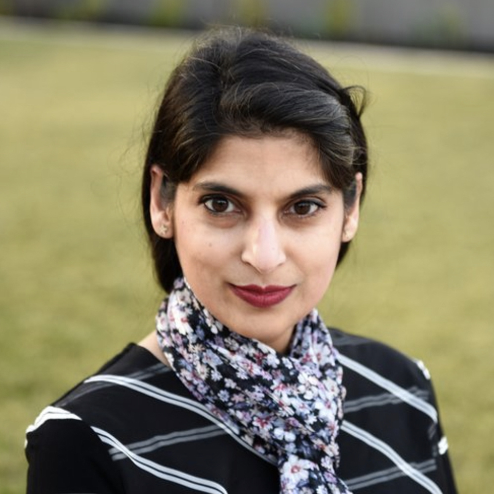 Photo: A portrait shot of Zainab Khan, a woman with dark hair. She wears red lipstick, a scarf, and a black and white shirt.