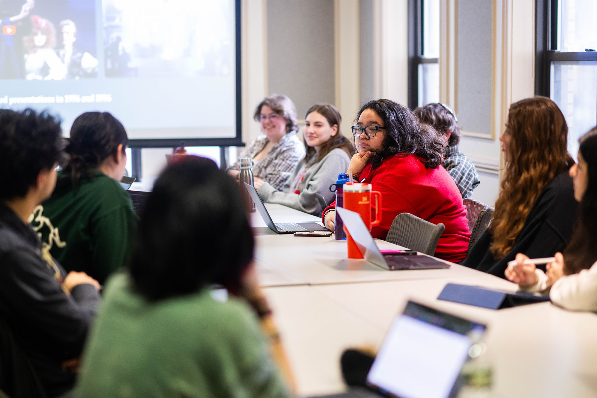 Photo: a classroom full of college students with a long table in the middle and a projector screen in the back of the room
