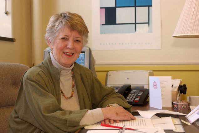 Photo: An older woman with a kind smile wearing a cardigan and an elegant necklace sits at a desk in an office environment