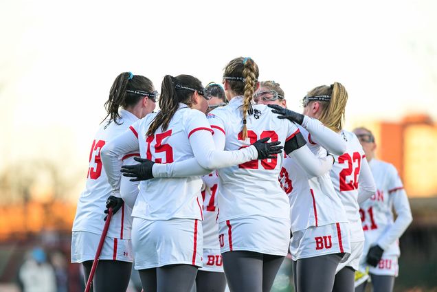 Photo: A picture of a woman's lacrosse team in a huddle. Their uniforms are white with red accents