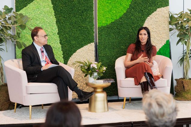 Photo: A picture of two people sitting in separate armchairs. There is a small table between them with flowers and books on it. They are dressed in formal attire