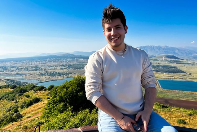 Photo: Michael Arellano, a young individual with short black hair wearing a white crew neck, sits casually on a stone wall overlooking a vast landscape. They smile for the photo.