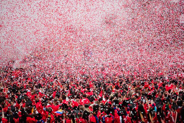 Photo: a field full of Boston University graduates wearing red gowns and black caps, with confetti falling all around