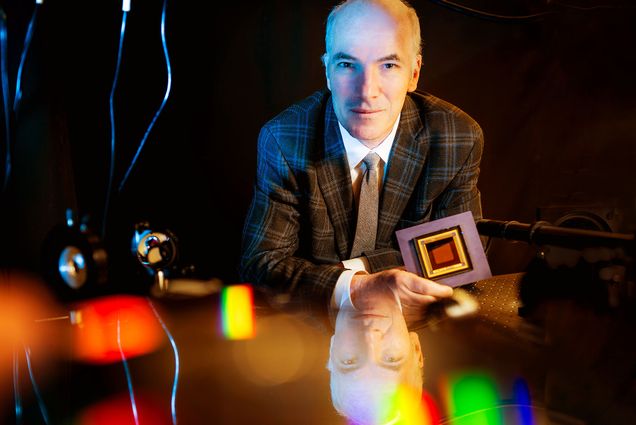 Photo: A picture of a man in a dark plaid suit looking at the camera. There is a reflection of his face on the table he is leaning against