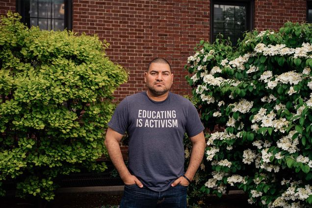 Photo: A man in a gray t shirt with text reading "Education is activism" stands in front of a brick wall with a stoic expression