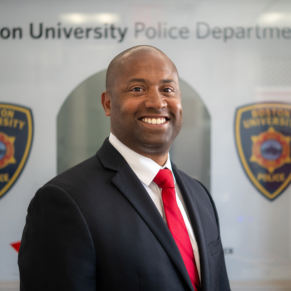 Photo: Boston University Police Chief Robert Lowe, a bald black man in a black suit with a red tie, standing in front of a wall displaying two emblems of the “Boston University Police Department.” The setting suggests an official or formal environment, possibly within a university police department context.