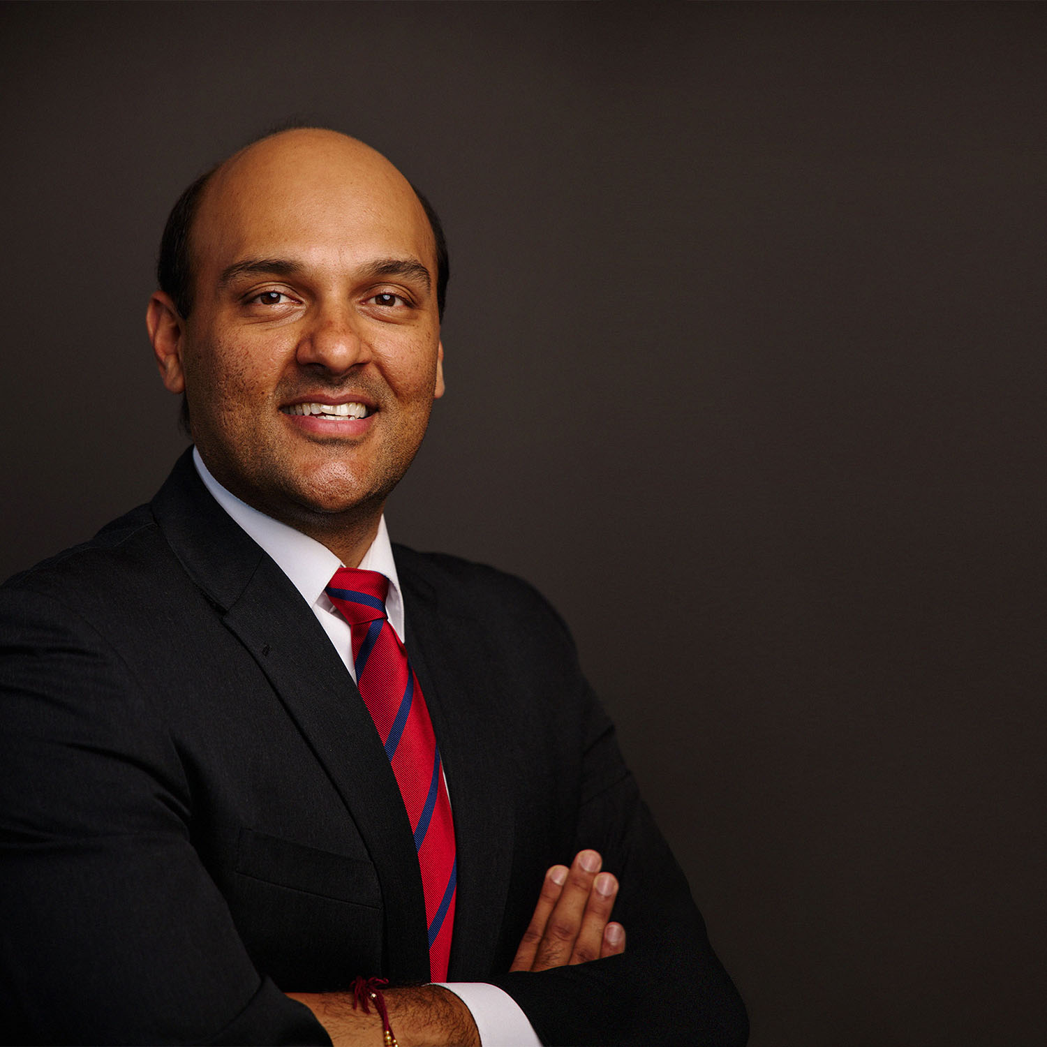 Photo: A headshot of a man in a black suit with a red striped tie and his arms crossed smiling for the camera