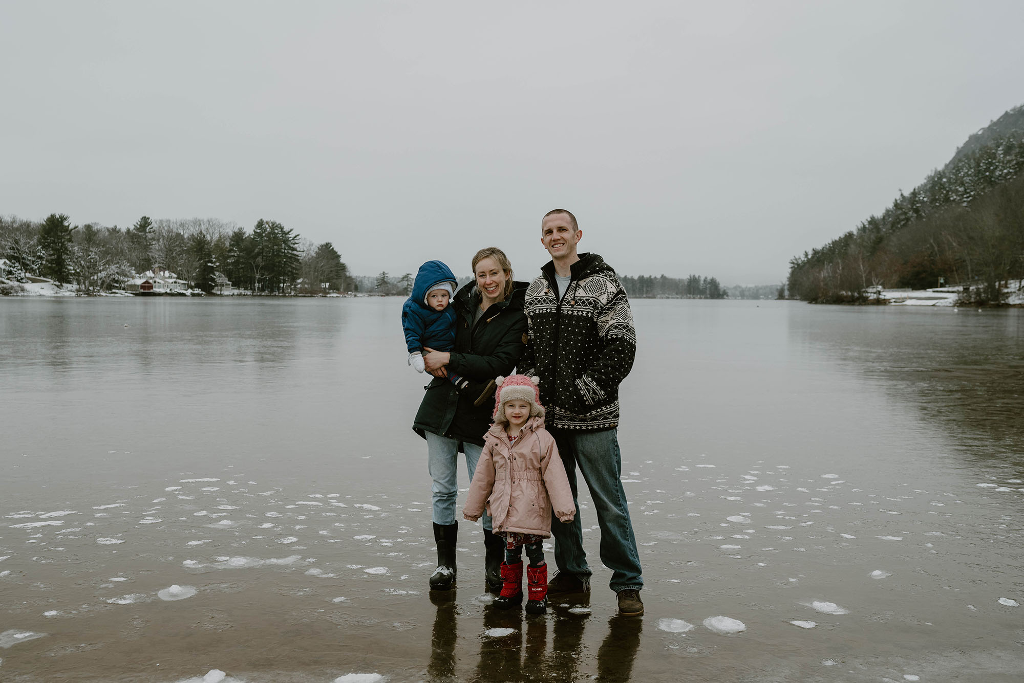 Photo: A white family standing on an iced over body of water in winter gear. The mother holds the youngest in her arms while the oldest stands in front of the mom and dad in her pink coat.