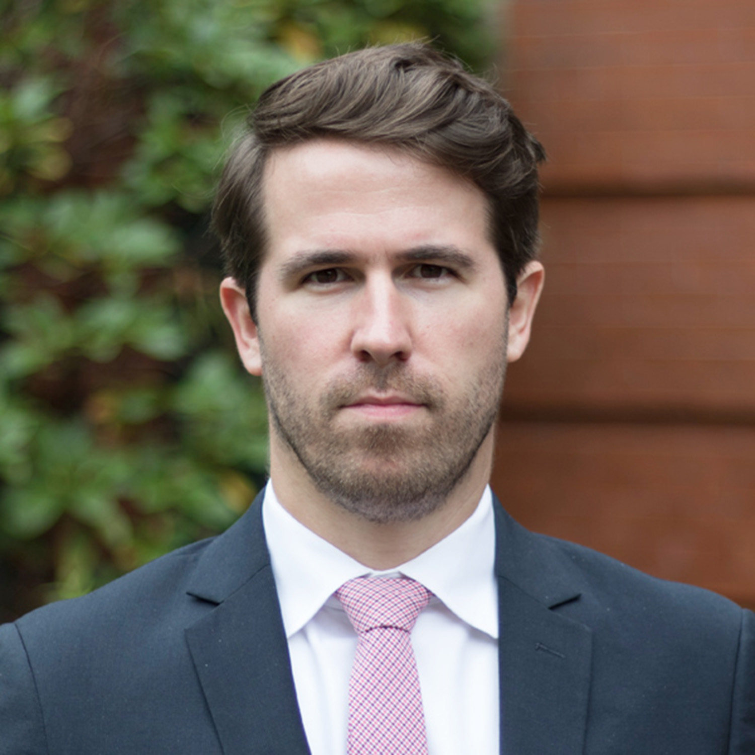 Photo: A headshot of a man with styled, brown hair looking at the camera. He is wearing a dark suit jacket with a white shirt and pink tie