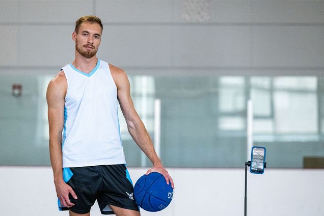 Photo: A muscular young man holding a blue basketball stands next to an iPhone on a tripod in a gym environment