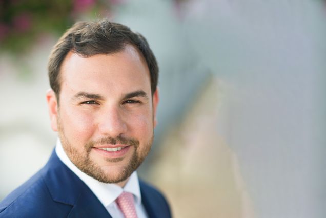 Photo: Formal headshot portrait of a white man with dark hair and beard, wearing a navy suit and white collared shirt.