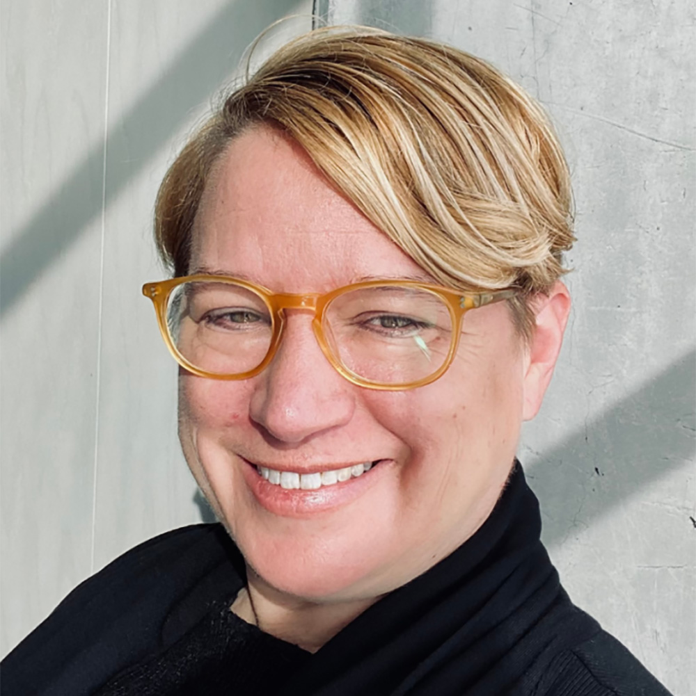 Photo: A portrait of a white woman with short blonde hair, orange frames, black shirt.