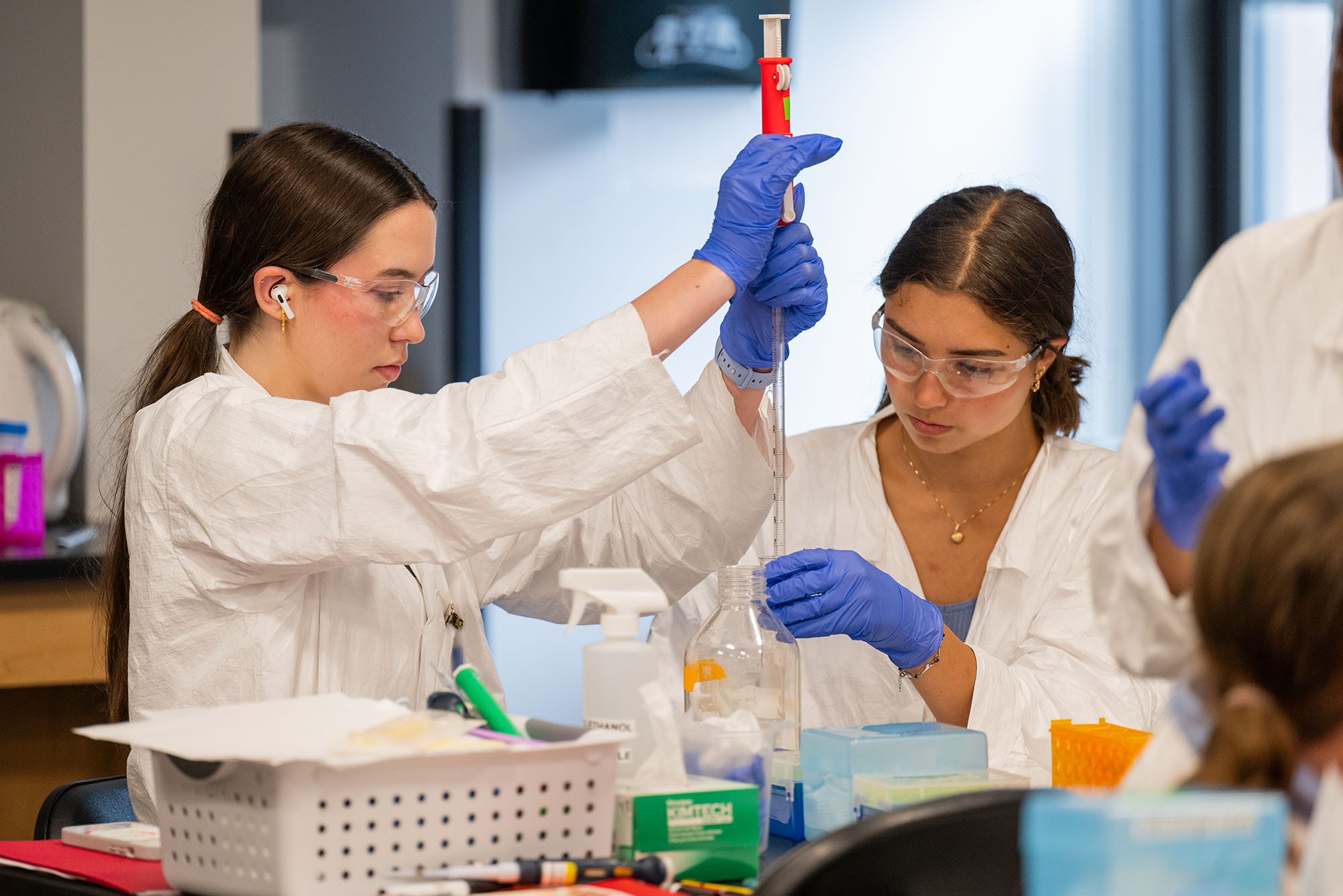 Photo: A picture of students in lab coats, gloves, and protective eyewear gathered around a lab table during an experiment
