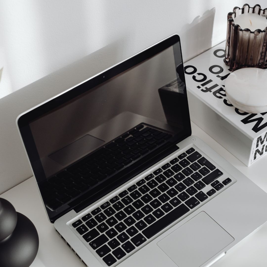 Photo: An open laptop on a white desk with a black screen, reflecting a window. The laptop is surrounded by various objects including a cup holder with pens, a book titled ‘LIFESTYLE’, and another object with text that is not fully visible. The scene suggests a tidy, modern workspace.