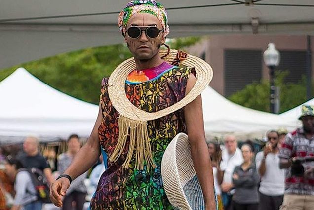 Photo: A picture of a person standing in front of a crowd while wearing colorful clothing with a fabric neck ring, hair scarf, and circular, black sunglasses. They are holding a woven basket