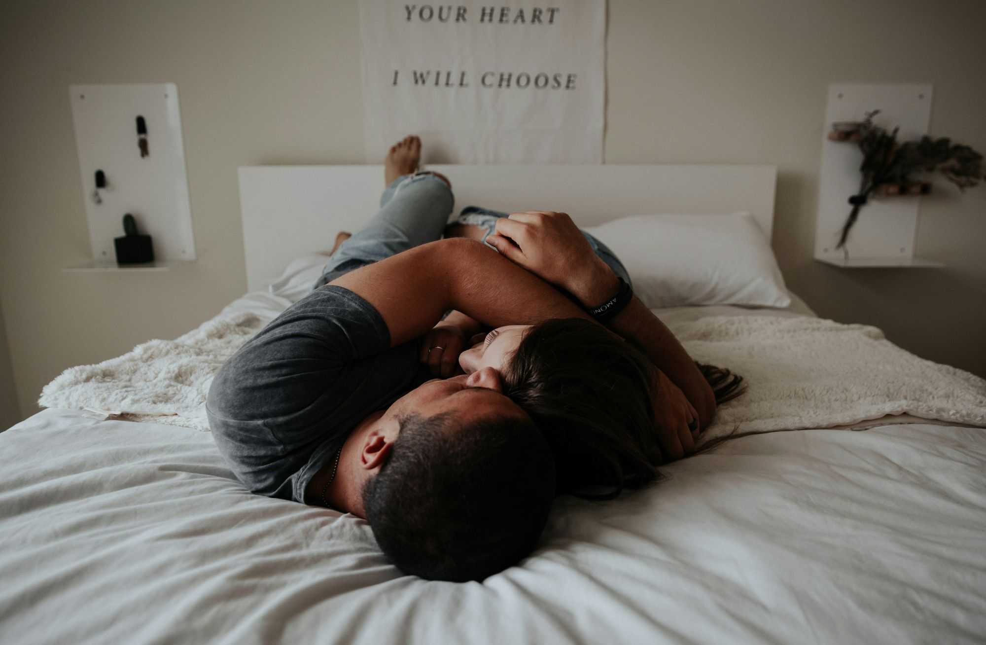 Photo: Two people, one with a tan skin tone, another with more fair skin tone, lay caressing on a bed with all white sheets. A motivational quote with the text ‘YOUR HEART I WILL CHOOSE’ written in bold, uppercase letters against a plain background.”