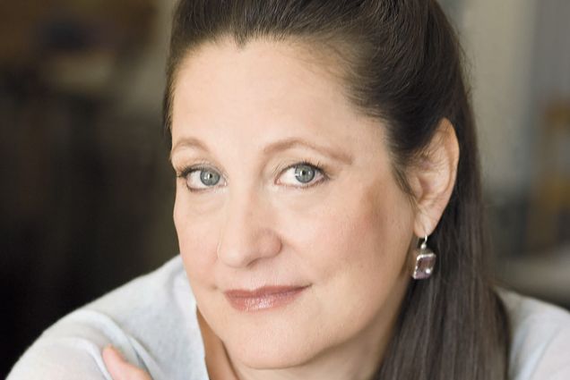Photo: A white woman with her brown hair pulled back. She wears silver jewelry and a white top.
