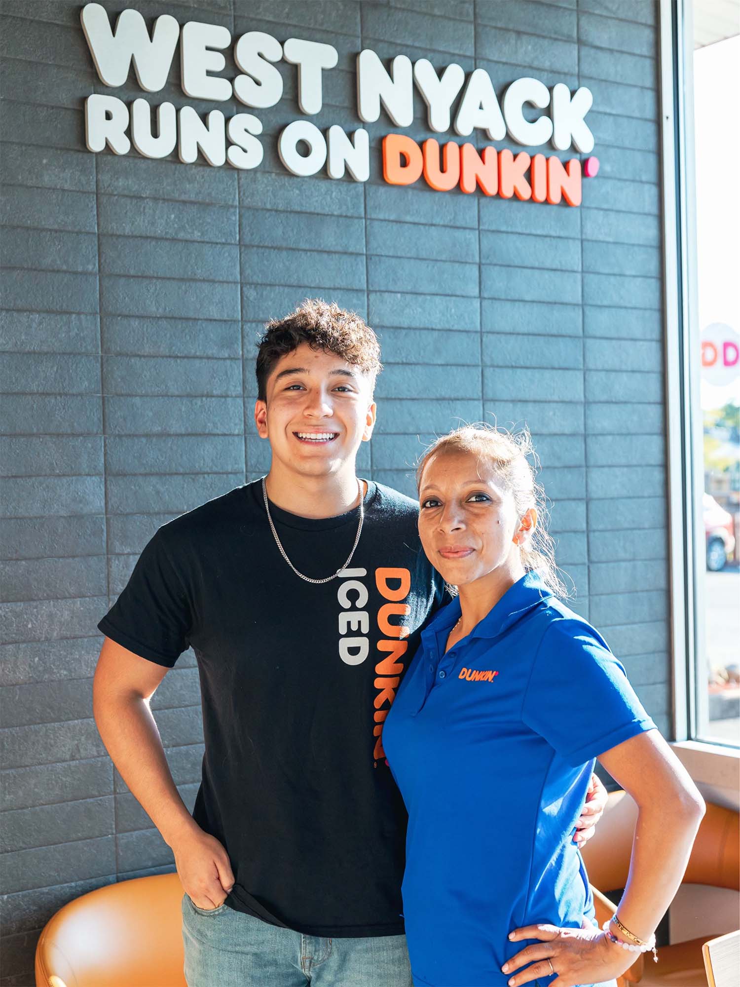 Photo: A young man smiling and posing with his mother. The man on the left is wearing a black tshirt and his mother has a blue shirt, both with the with the Dunkin' orange logo.